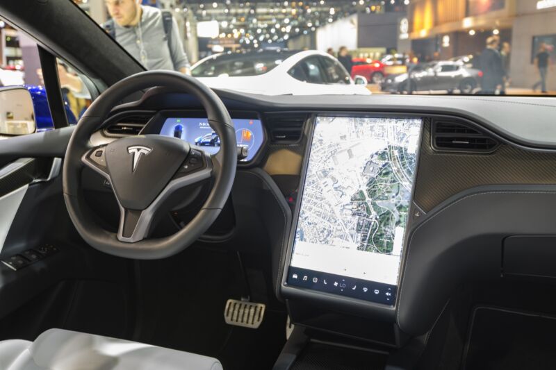The interior of a Tesla Model X SUV. A large touch screen next to the steering wheel displays a map.